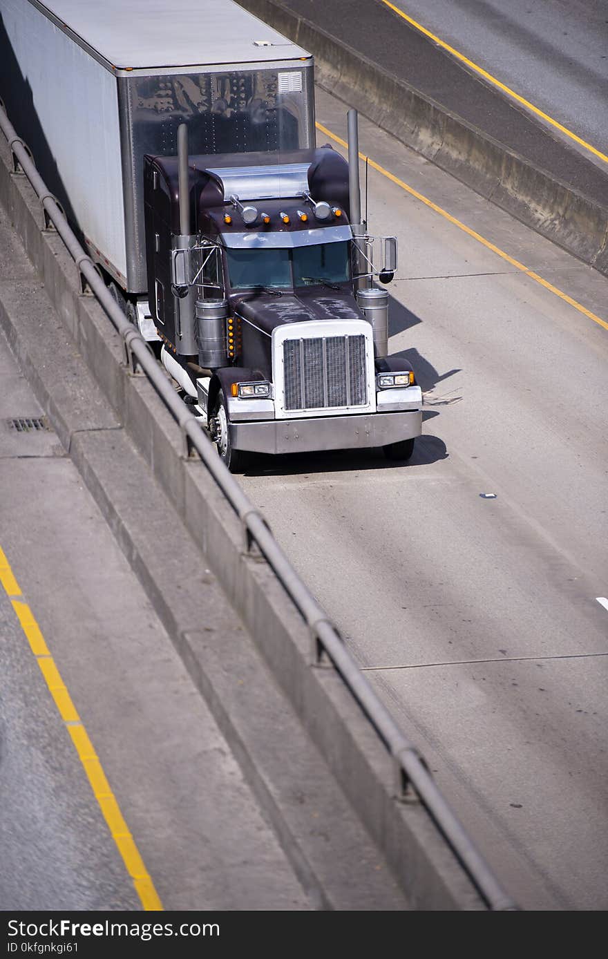 Dark old classic big rig semi truck idol with chrome accessories and tall exhaust pipes transporting cargo at dry van semi trailer with stainless steel wall on divided multiline highway. Dark old classic big rig semi truck idol with chrome accessories and tall exhaust pipes transporting cargo at dry van semi trailer with stainless steel wall on divided multiline highway.