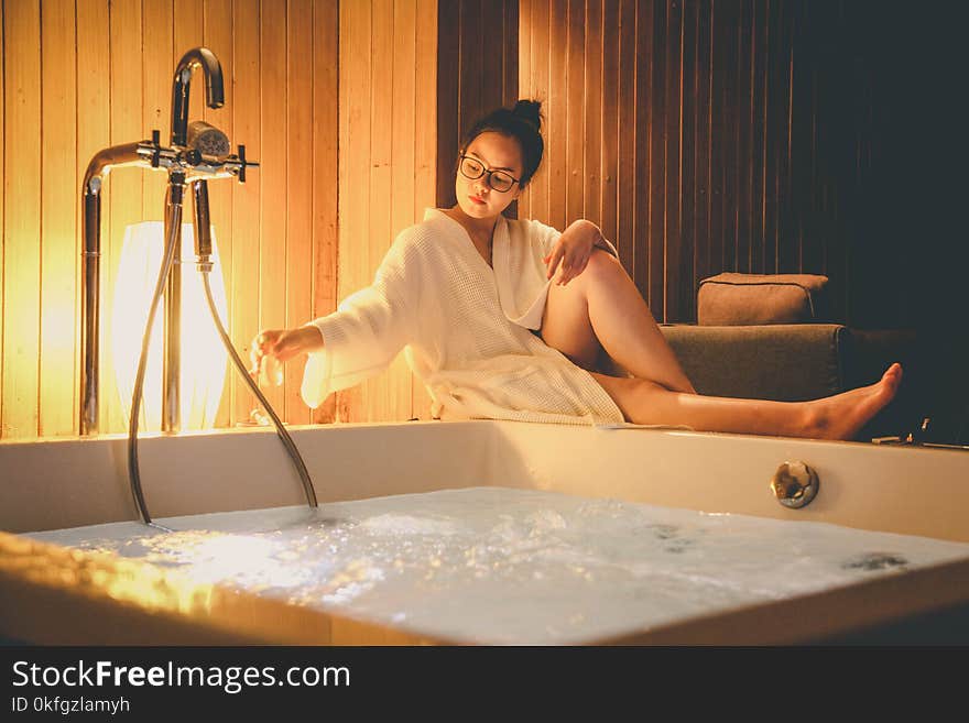 Woman Wearing White Bathrobe Sitting Beside White Bathtub Filled With Water