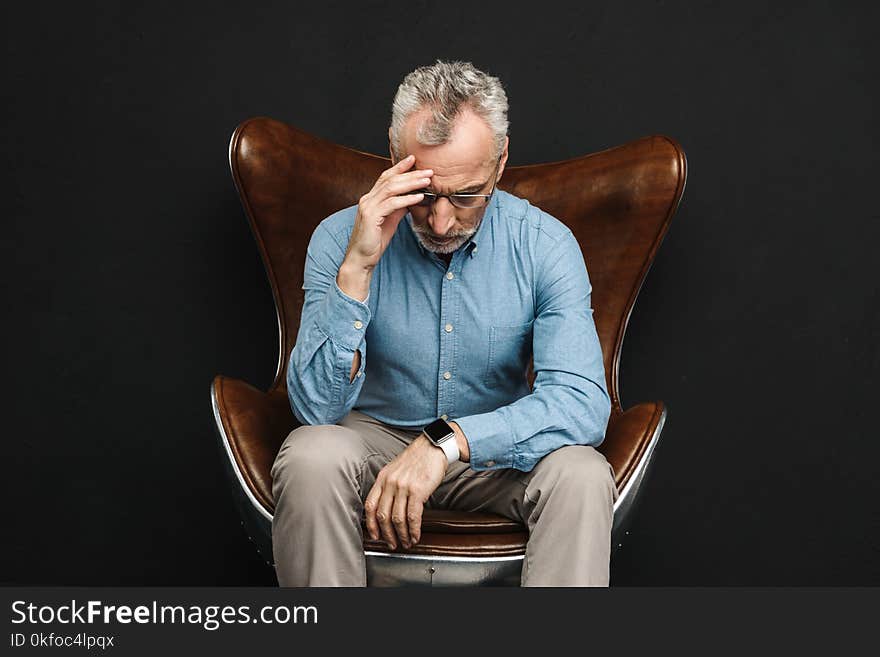 Image of intelligent gentleman 50s having grey hair and beard in glasses sitting on businesslike armchair with face downward isolated over black background