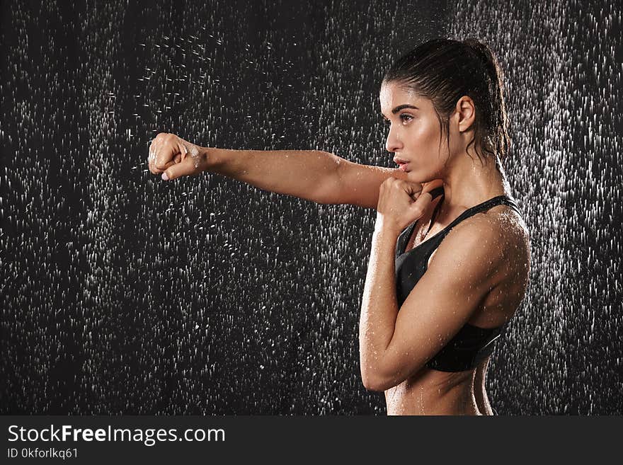 Photo in profile of focused athletic woman wearing black sportive bra punchning with clenched fist isolated over rain drops background