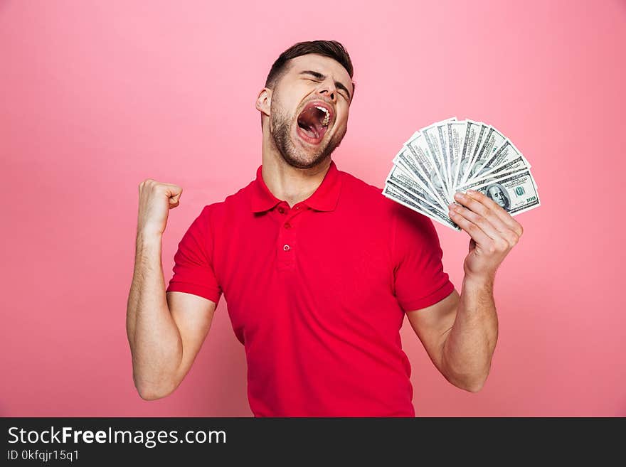 Portrait of a satisfied young man holding money banknotes and celebrating success isolated over pink background