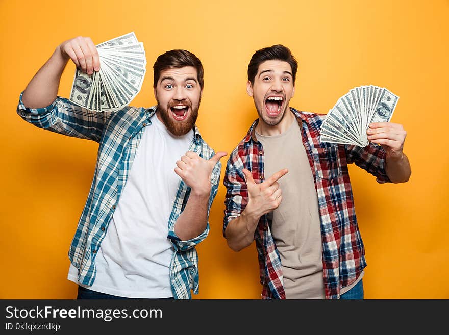 Portrait of a two excited oung men showing money banknotes and pointing fingers isolated over yellow background