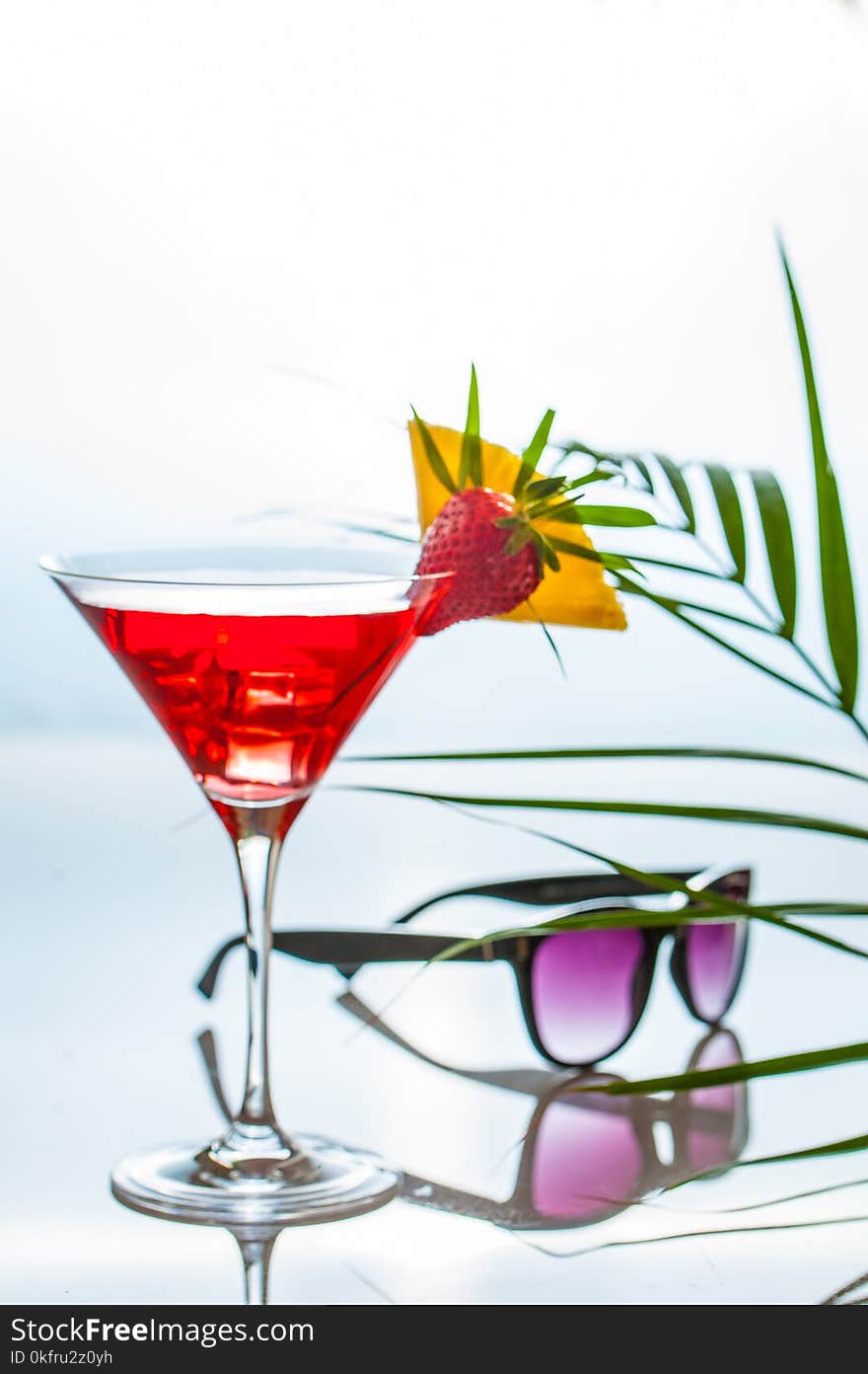 Colorful tropical cocktail in a martini glass decorated by slices of tropical fruits and a leaves on a reflecting table with sunglasses and palm tree leaves ona side. Negative space.