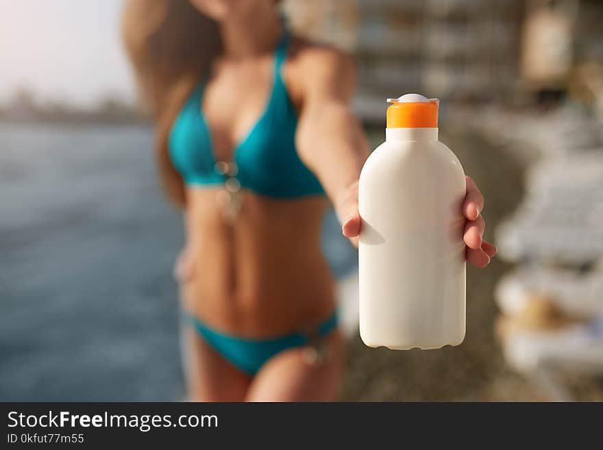 Happy woman shows the suntan or sunscreen cream white bottle over the blurred beach background. Tanned slim girl wearing