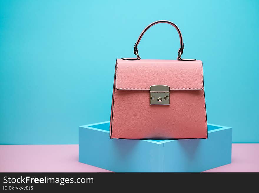 Coral leather female bag on the blue decorative stand on the pink surface on the cyan background in the studio. Closeup. Horizontal. Coral leather female bag on the blue decorative stand on the pink surface on the cyan background in the studio. Closeup. Horizontal.