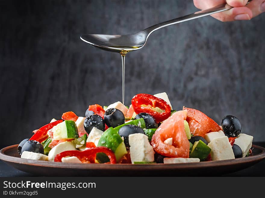 Woman`s hand is pouring a Greek salad with olive oil from a spoon on dark background. Woman`s hand is pouring a Greek salad with olive oil from a spoon on dark background