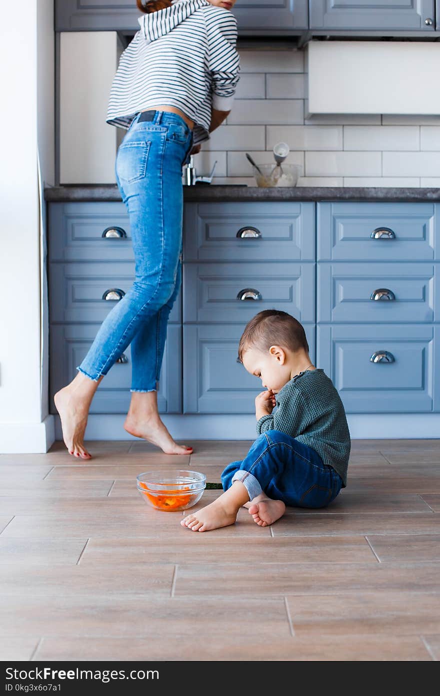 A cute little boy is sitting on the kitchen floor with his mother in the background