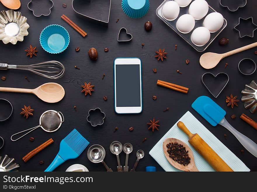 Baking background with eggs and kitchen tools: rolling pin, wooden spoons, whisk, sieve, bakeware, shape cookie cutter and mobile phone mockup on dark wooden background. Horizontal orientation with copyspace, top view. Baking background with eggs and kitchen tools: rolling pin, wooden spoons, whisk, sieve, bakeware, shape cookie cutter and mobile phone mockup on dark wooden background. Horizontal orientation with copyspace, top view.