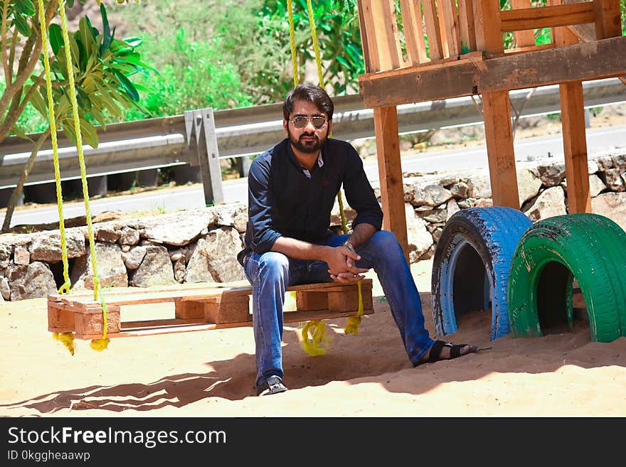 Man Wearing Black Dress Shirt and Blue Jeans Sitting on Wooden Pallet Swing