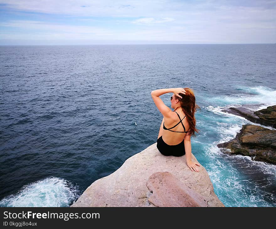 Woman in Black Swimwear Set Sitting on Top of Gray Rocky Cliff Facing Body of Water