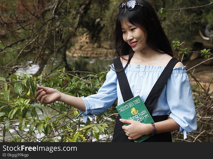 Woman Wearing Blue Off-shoulder Blouse Holding Book Next to Green Leaf Plant