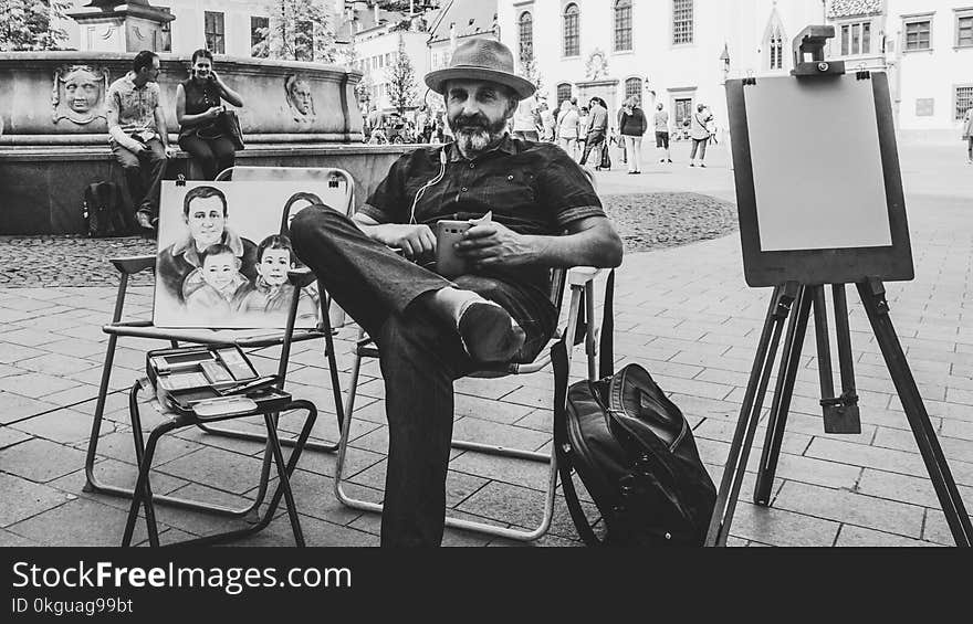 Grayscale Photo of Man Wearing Black Dress Shirt With Hat Sitting on Folding Armchair Beside Family Sketch