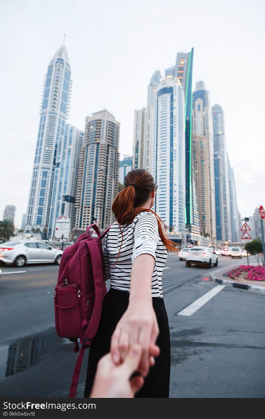 A beautiful girl with a backpack leads behind her and holds her hand against the background of tall buildings