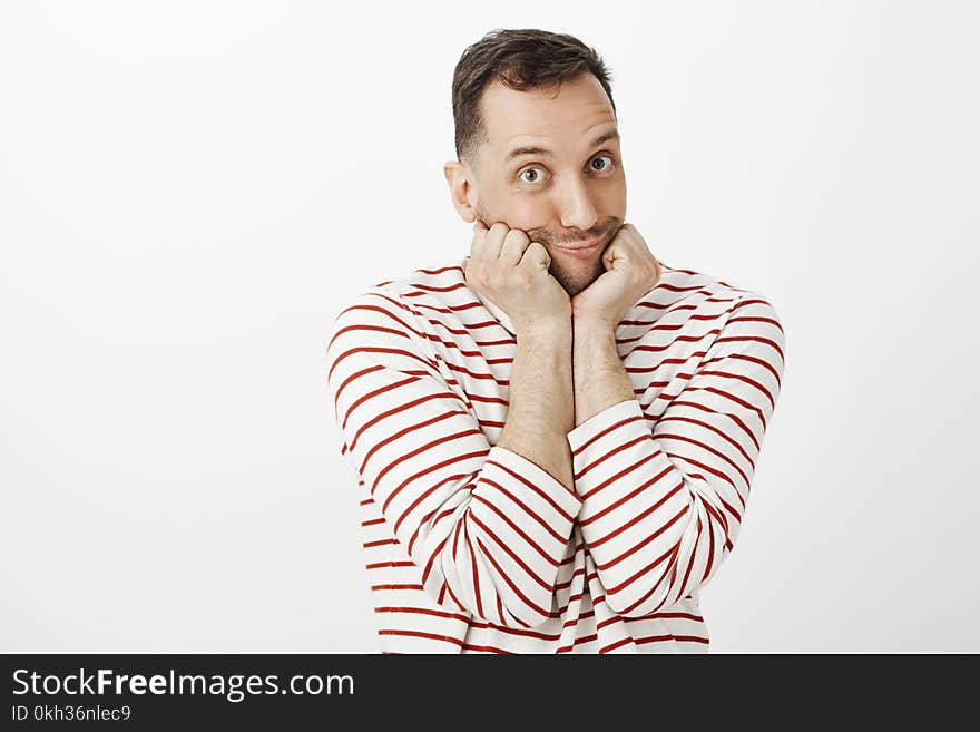Indoor shot of cute emotive gay man in striped pullover, leaning face on palms and making tender duck face with pouting lips, flirting with boyfriend and standing against gray background. Copy space