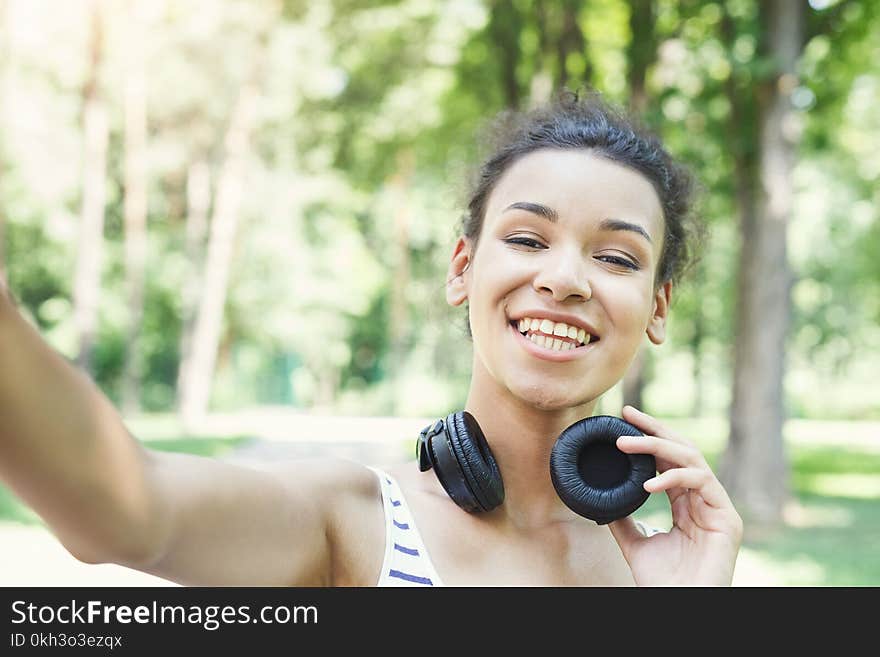 Happy african-american woman taking selfie in sunny summer park, while listening music, relaxing from jog training, copy space. Happy african-american woman taking selfie in sunny summer park, while listening music, relaxing from jog training, copy space