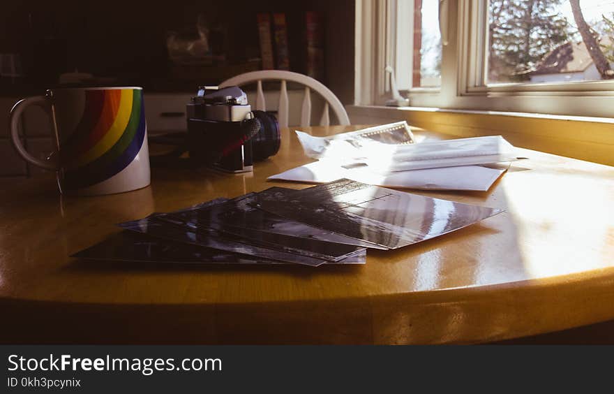 This vintage look photograph is warm with morning sun coming in the window. An old SLR camera from the 70`s sits next to a rainbow coffee mug. Black & white photographic prints are scattered across the table, along with the envelope from the processing lab & the pack of 35mm film negatives. This vintage look photograph is warm with morning sun coming in the window. An old SLR camera from the 70`s sits next to a rainbow coffee mug. Black & white photographic prints are scattered across the table, along with the envelope from the processing lab & the pack of 35mm film negatives.