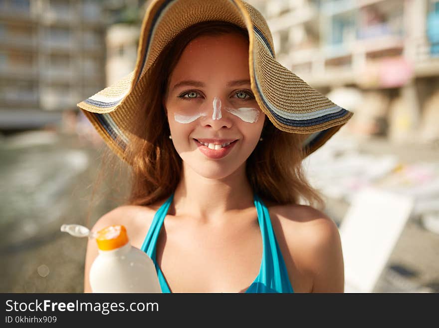 Suntan lotion. Woman applying sunscreen solar cream on face. Beautiful happy cute girl puts suntan cream from plastic container bottle on her nose and cheecks. Skincare concept. Female in straw hat and bikini on beach.