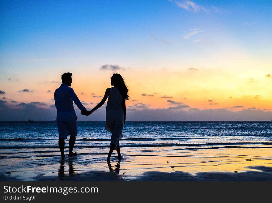 Man and Woman Holding Hands Walking on Seashore during Sunrise