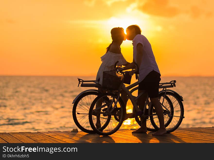 Photography of Man Wearing White T-shirt Kissing a Woman While Holding Bicycle on River Dock during Sunset