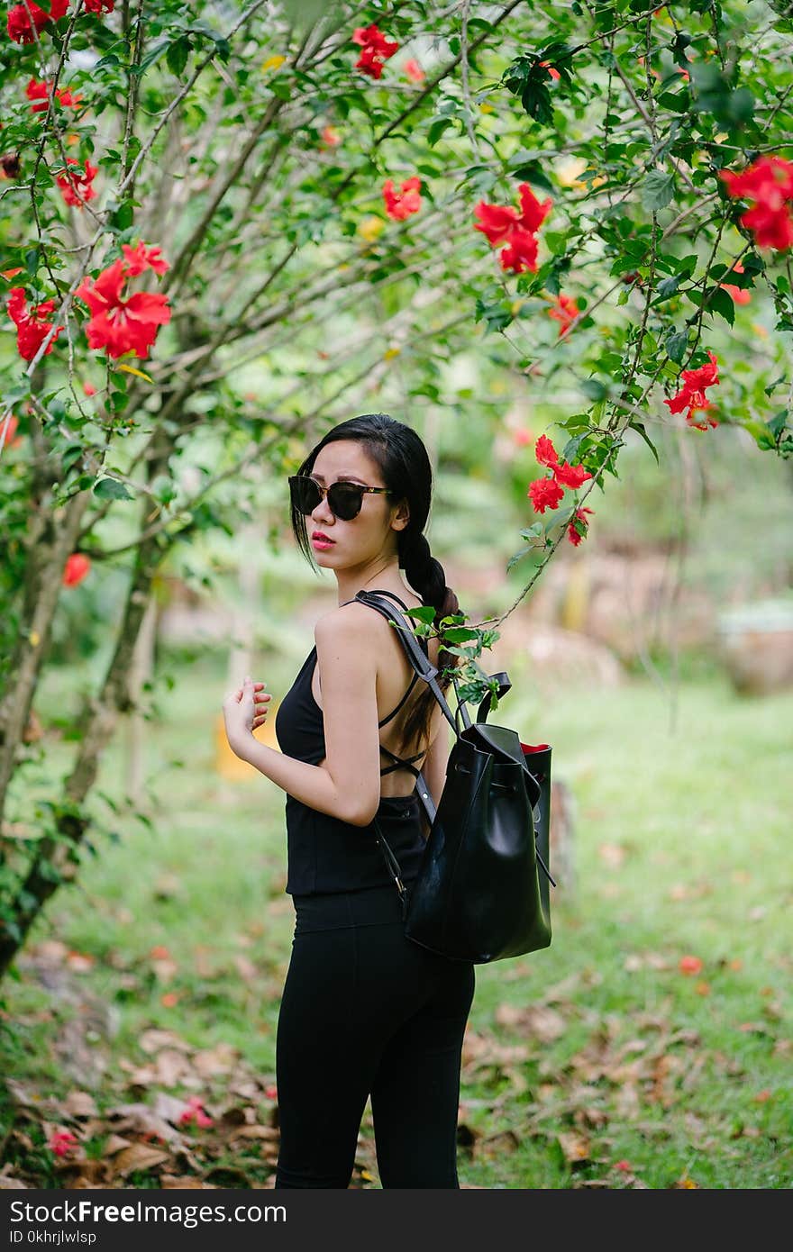 Woman Looking over Her Shoulder Wearing Black Sleeveless Top and Black Jeans Under a Red Hibiscus Plant at Daytime