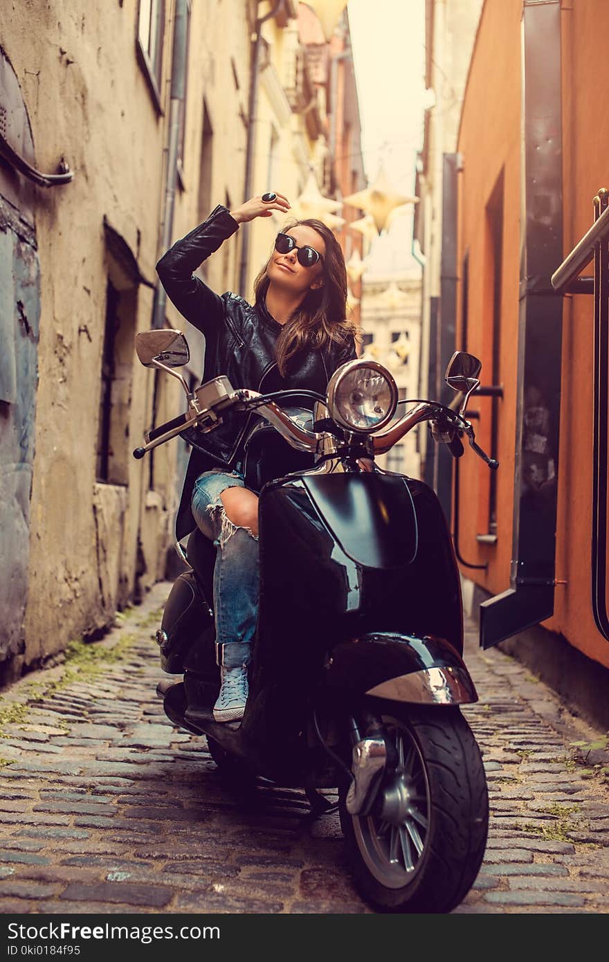 Attractive brunette female in leather jacket sitting on a motor scooter on a street in old town. Attractive brunette female in leather jacket sitting on a motor scooter on a street in old town.