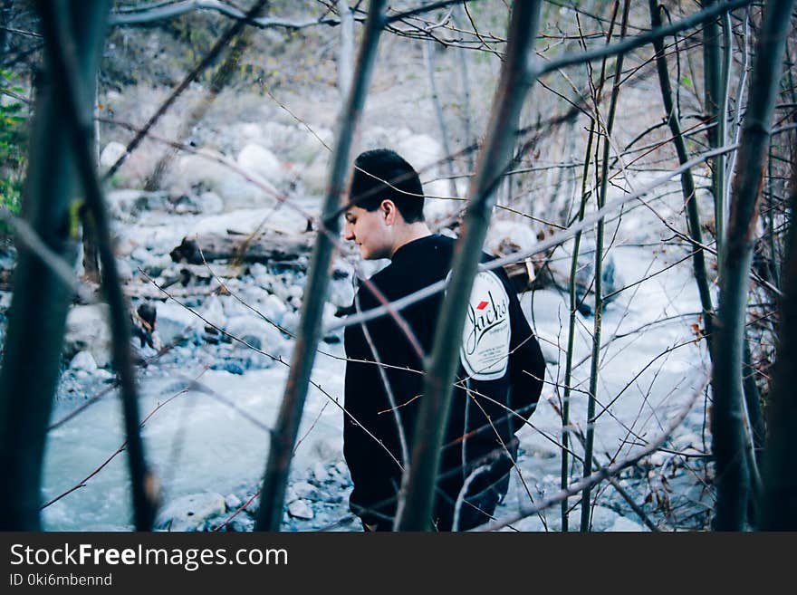 Man Wearing Black Sweatshirt Standing Beside River and Bare Trees