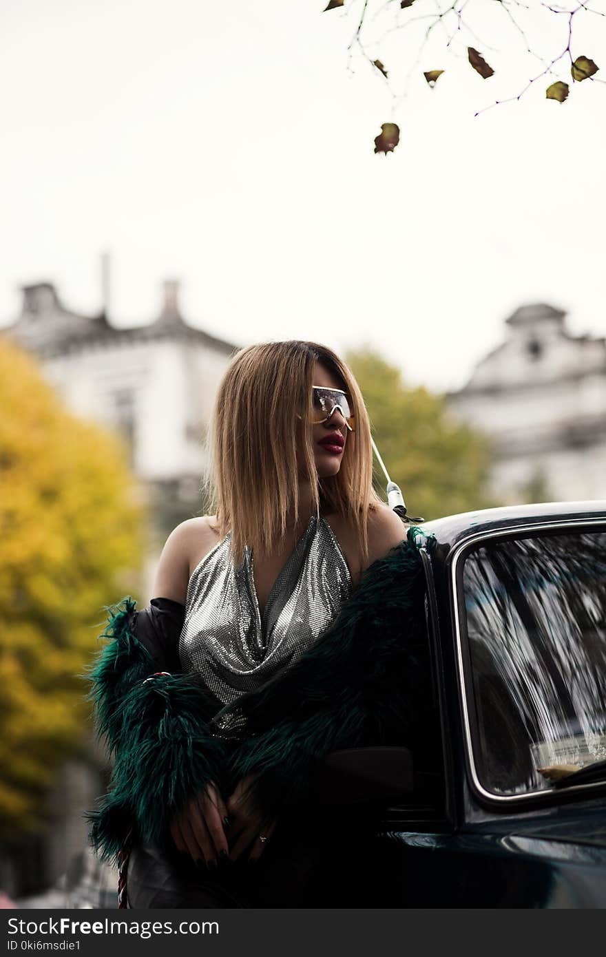 Selective Focus Photography of Woman Wearing Silver V-neck Sleeveless Top Leaning on Black Car