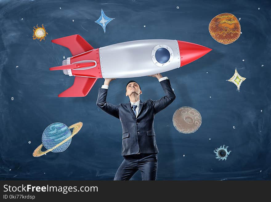 A businessman holds a model of a retro rocked above his head near drawings of planets and stars. Bring dreams to live. Business goals. Strength for success.