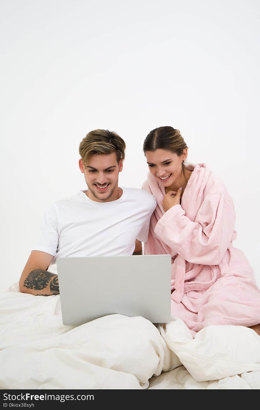 Man in White Crew-neck Shirt Sitting on Bed Beside Woman in Pink Bathrobe Looking at Laptop Computer