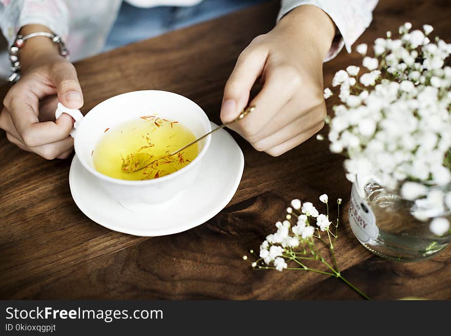 Person Holding Silver Spoon on White Ceramic Teacup With Yellow Liquid Inside