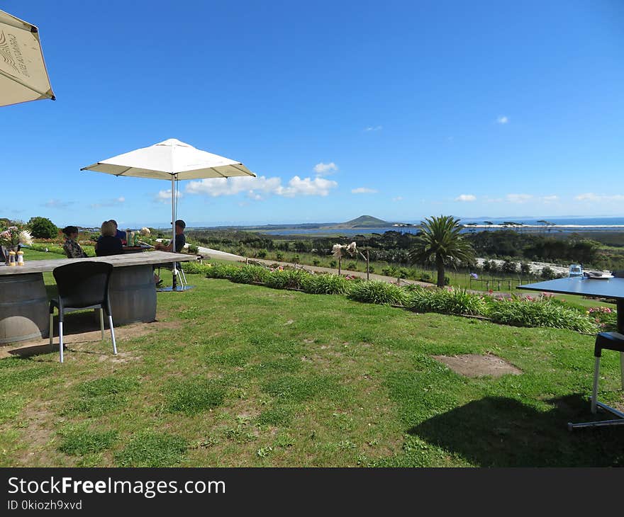 People Sitting in Front of Patio Table With Parasol Placed on Hillside With View of Body of Water and Mountain