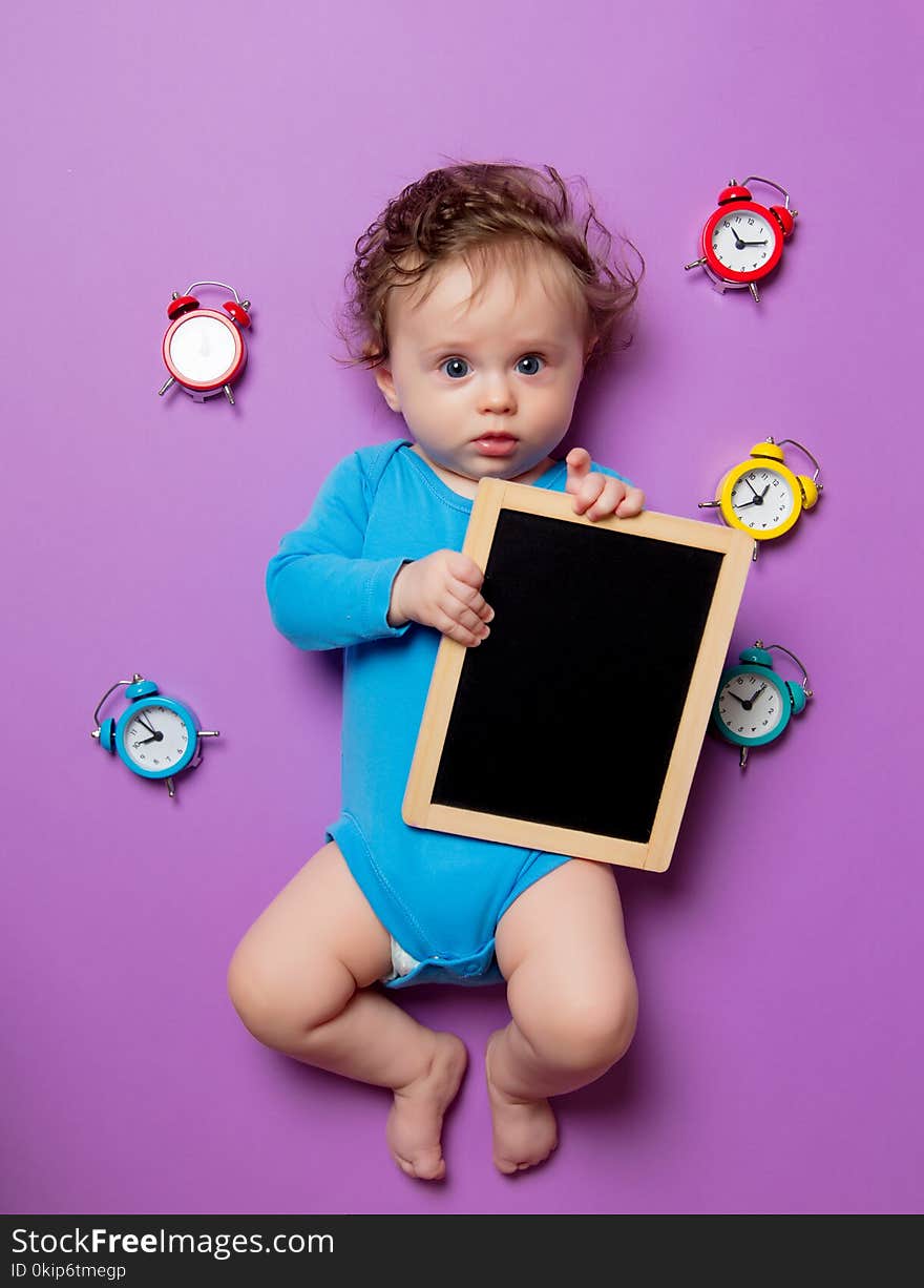 Little infant baby with alarm clock and blackboard lying down on purple background
