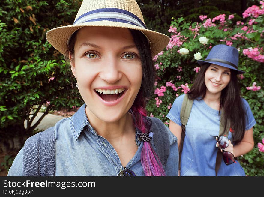 Two young beautiful brunette girls in blue jeans dresses and hats are laughing, posing in the garden with blooming roses on the fisheye lens, summer, sunny day, good mood, laughter and fun. Two young beautiful brunette girls in blue jeans dresses and hats are laughing, posing in the garden with blooming roses on the fisheye lens, summer, sunny day, good mood, laughter and fun.