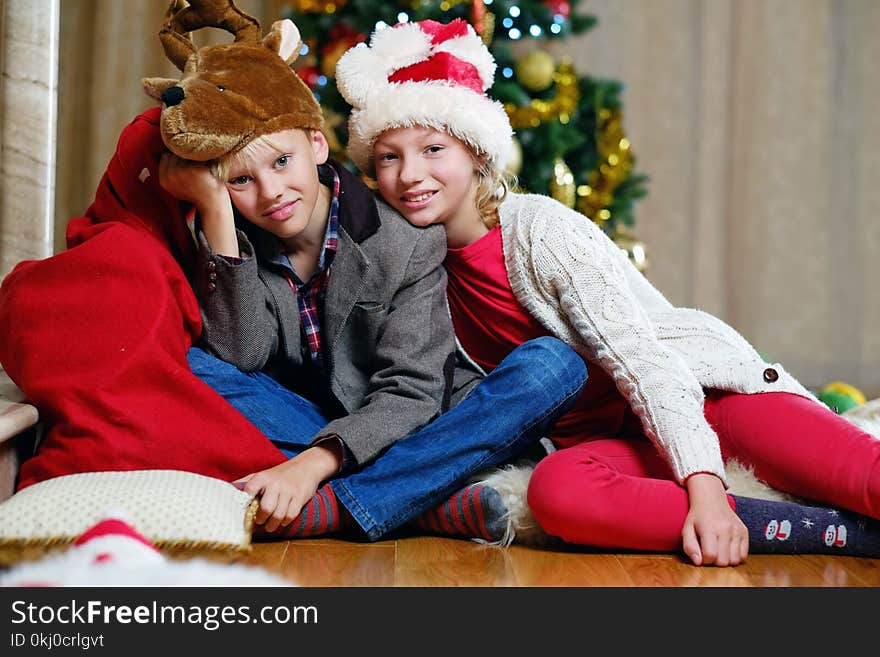 Smiling teenage girl and boy sitting on the floor with a Christmas tree in background.