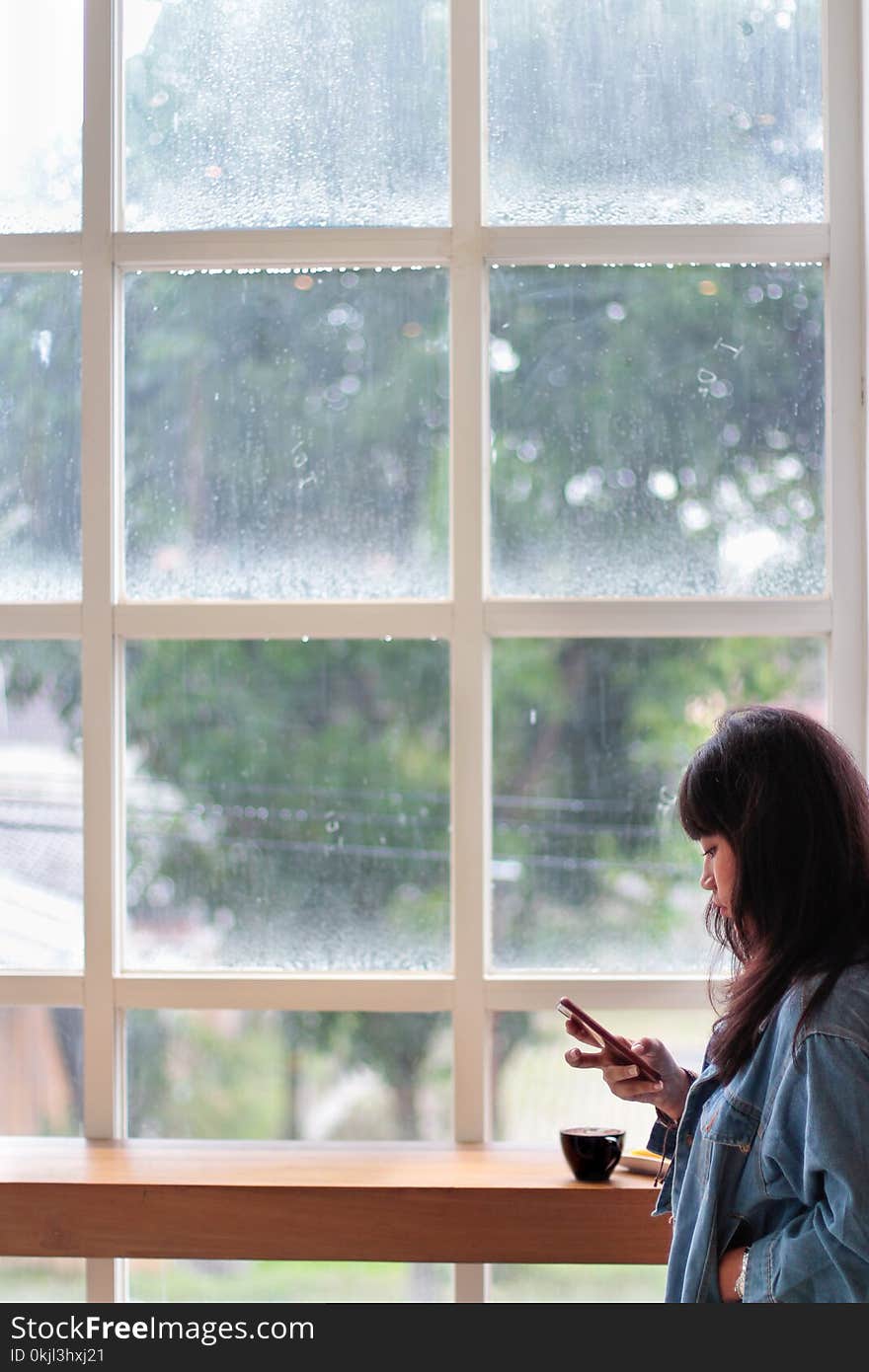 Woman Wearing Blue Denim Jacket Holding Smartphone Standing Beside Clear Glass Window