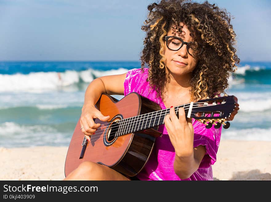 Woman Wearing Purple Shirt Playing Brown Classical Guitar While Sitting Near Shoreline With Water Splashing Background in Daytime