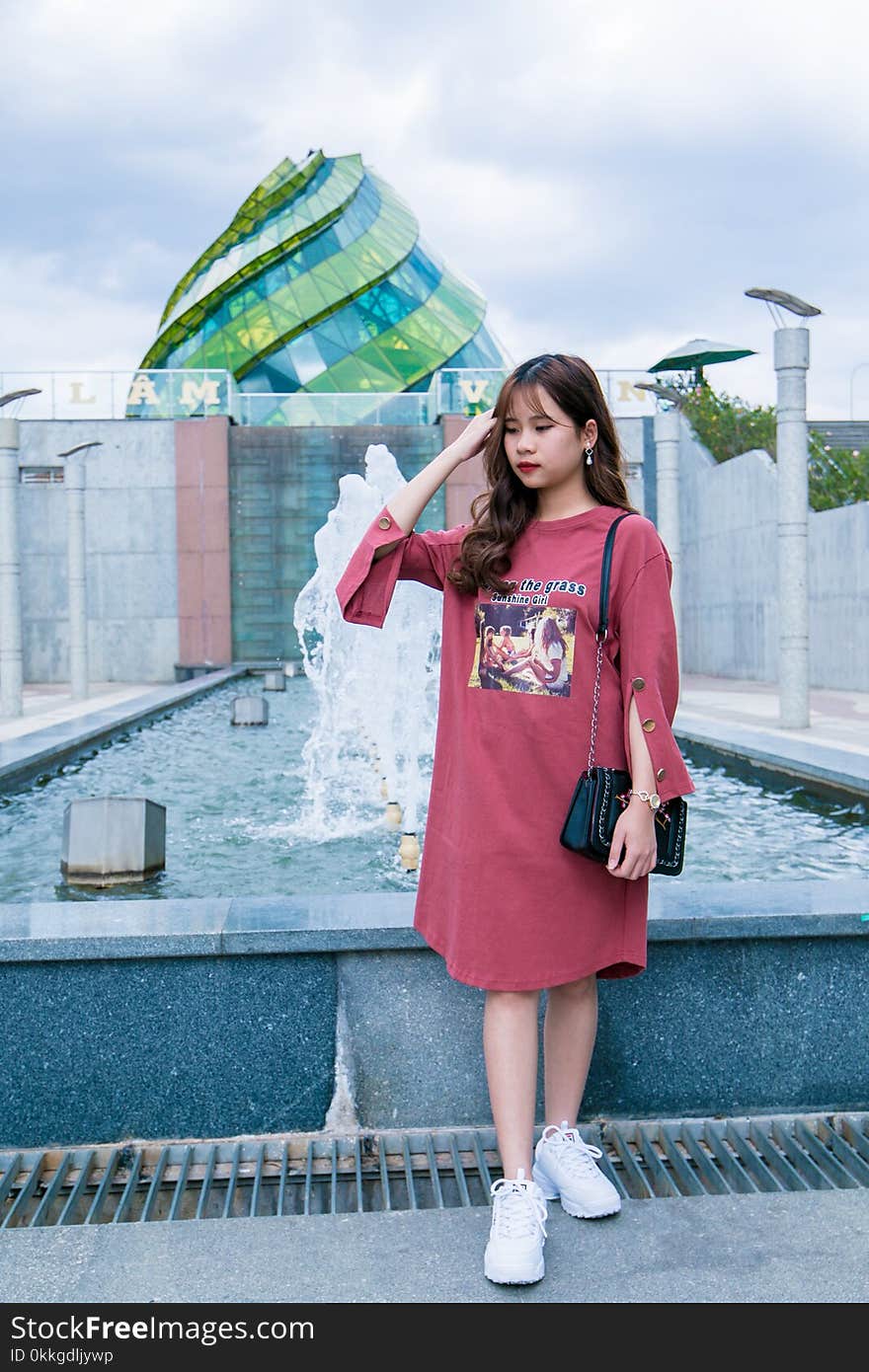 Woman Wearing Maroon Crew-neck Long-sleeved Dress Standing Next to Fountain Under Cloudy Sky