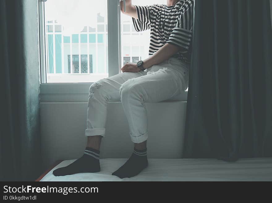 Woman Sitting on Window Stepping on White Bed Inside Well Lighted Room