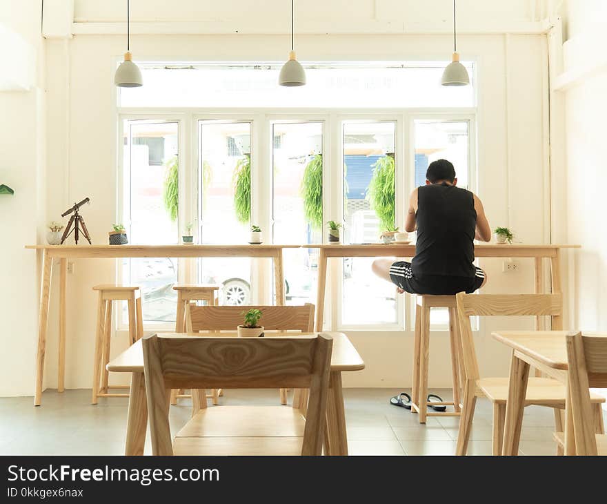 Man in Black Sleeveless Shirt Sitting on Wooden High Chair Beside Wooden Tables Near Window