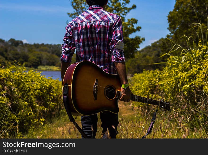 Man in Pink, Black, and White Plaid Dress Shirt Holding a Guitar Near Green Bush