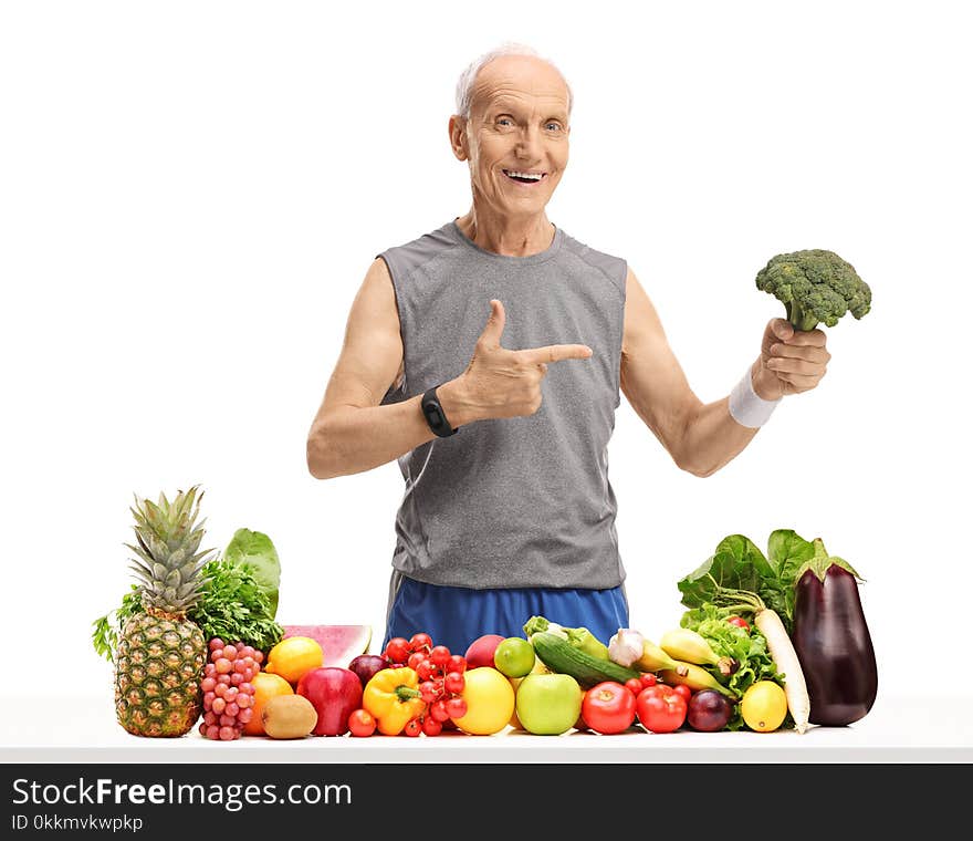 Elderly man behind a table with fruit and vegetables holding broccoli and pointing isolated on white background