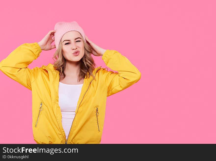 Crazy beautiful trendy girl in bright yellow jacket and pink beanie hat puckering lips. Attractive cool young woman fashion portrait over pastel pink background. Crazy beautiful trendy girl in bright yellow jacket and pink beanie hat puckering lips. Attractive cool young woman fashion portrait over pastel pink background.