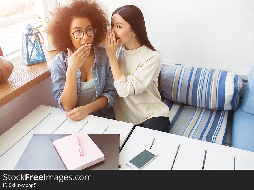 Girl in white sweater is whispering something on her friend`s ear. The afro american girl is wondering. She is amazed and putting the hand over the mouth