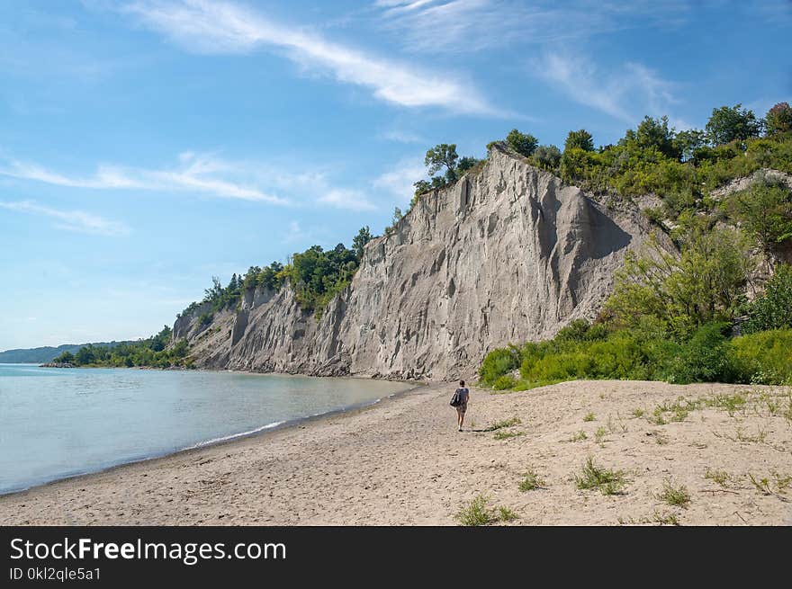 Woman Walks on Brown Seashore Near Cliff With Green Trees Under Blue and White Sky