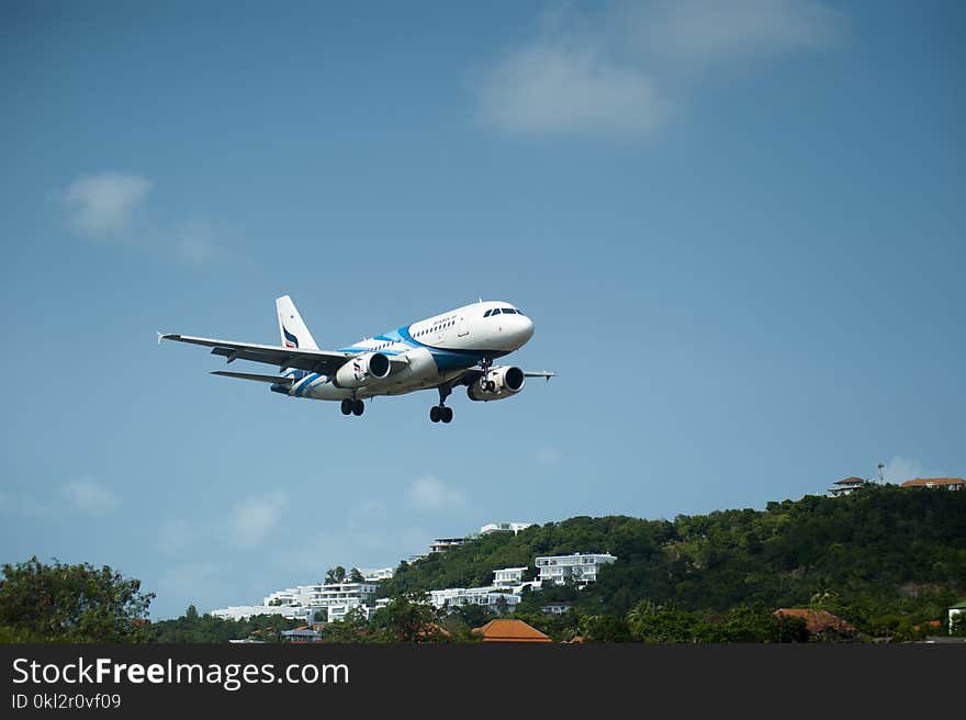 White and Blue Passenger Plane Passing Above Green Tree Covered Hill