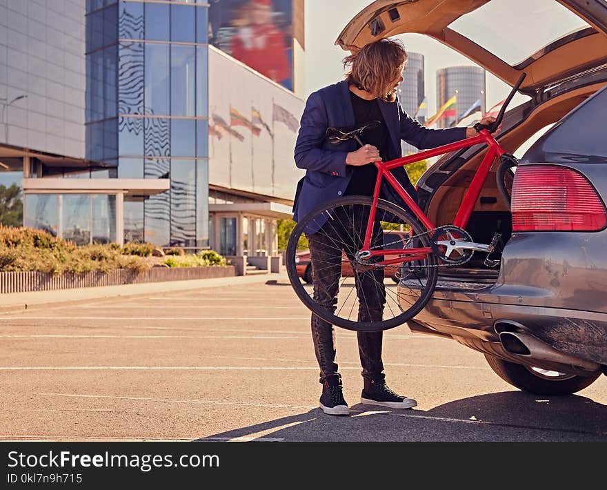 Bearded blond male with long hair puts fixed bicycle in the car`s trunk. Bearded blond male with long hair puts fixed bicycle in the car`s trunk.