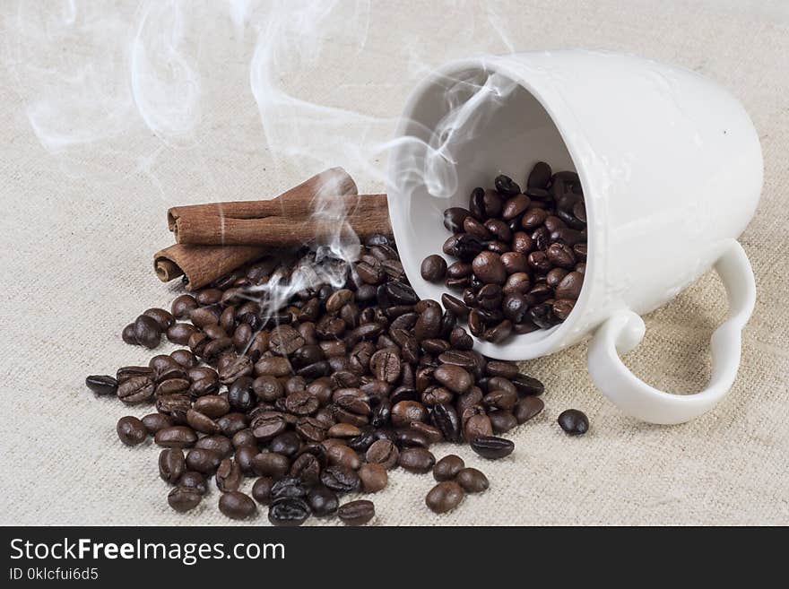 Black Fresh roasted coffee bean smoke and spilled from white coffee cup and cinnamon on retro fabric tablecloth on desk. Close up, selective focus. Retro food and drink concept image. Black Fresh roasted coffee bean smoke and spilled from white coffee cup and cinnamon on retro fabric tablecloth on desk. Close up, selective focus. Retro food and drink concept image.