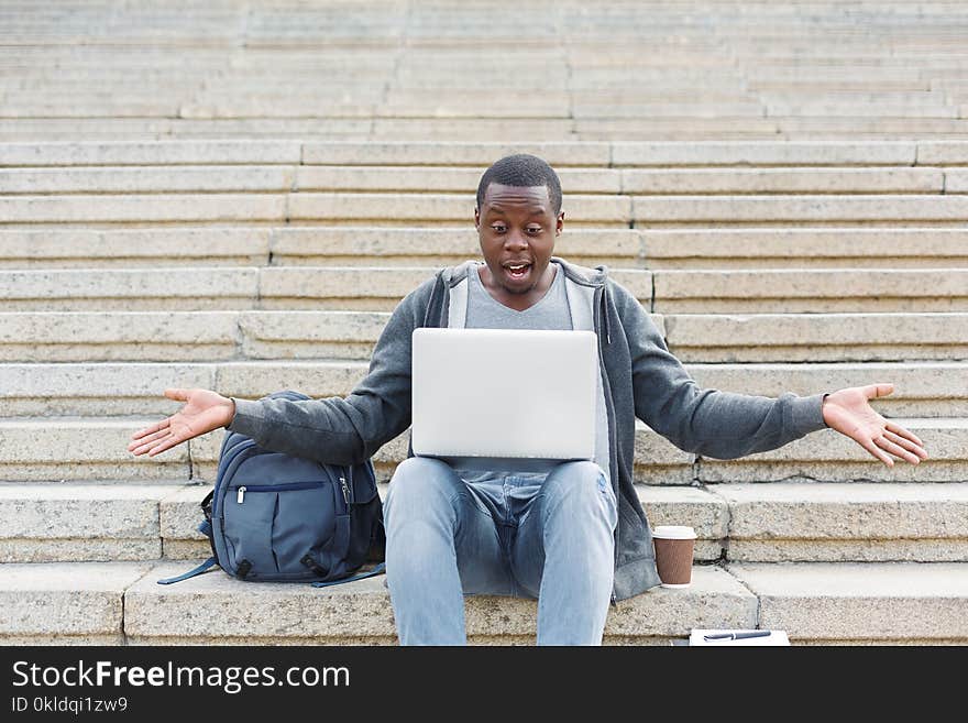 Desperate student sitting on stairs and working on laptop outdoors in university campus. Shocked african-american man raised his hands, copy space. Desperate student sitting on stairs and working on laptop outdoors in university campus. Shocked african-american man raised his hands, copy space