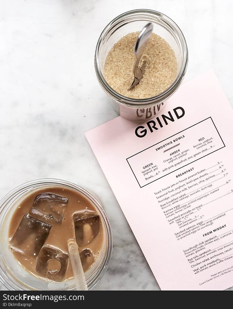 Clear Drinking Glass Filled With Tea and Ice Cubes With Clear Plastic Straw Beside Clear Glass Canister Filled With Brown Sugar