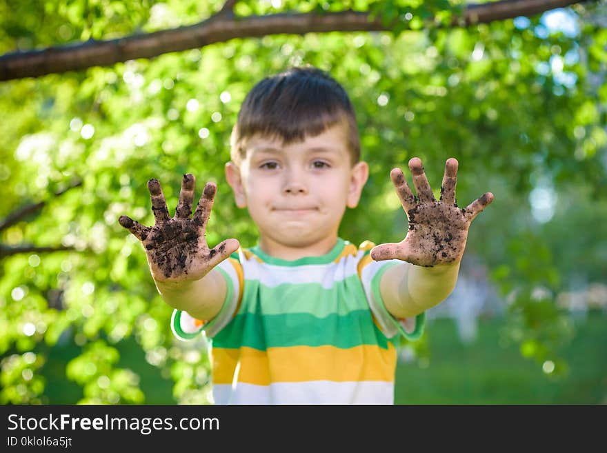 A young Caucasian boy showing off his dirty hands after playing in dirt and sand outdoors sunny spring or summer evening on blossom trees background. happy childhood friendship concept.